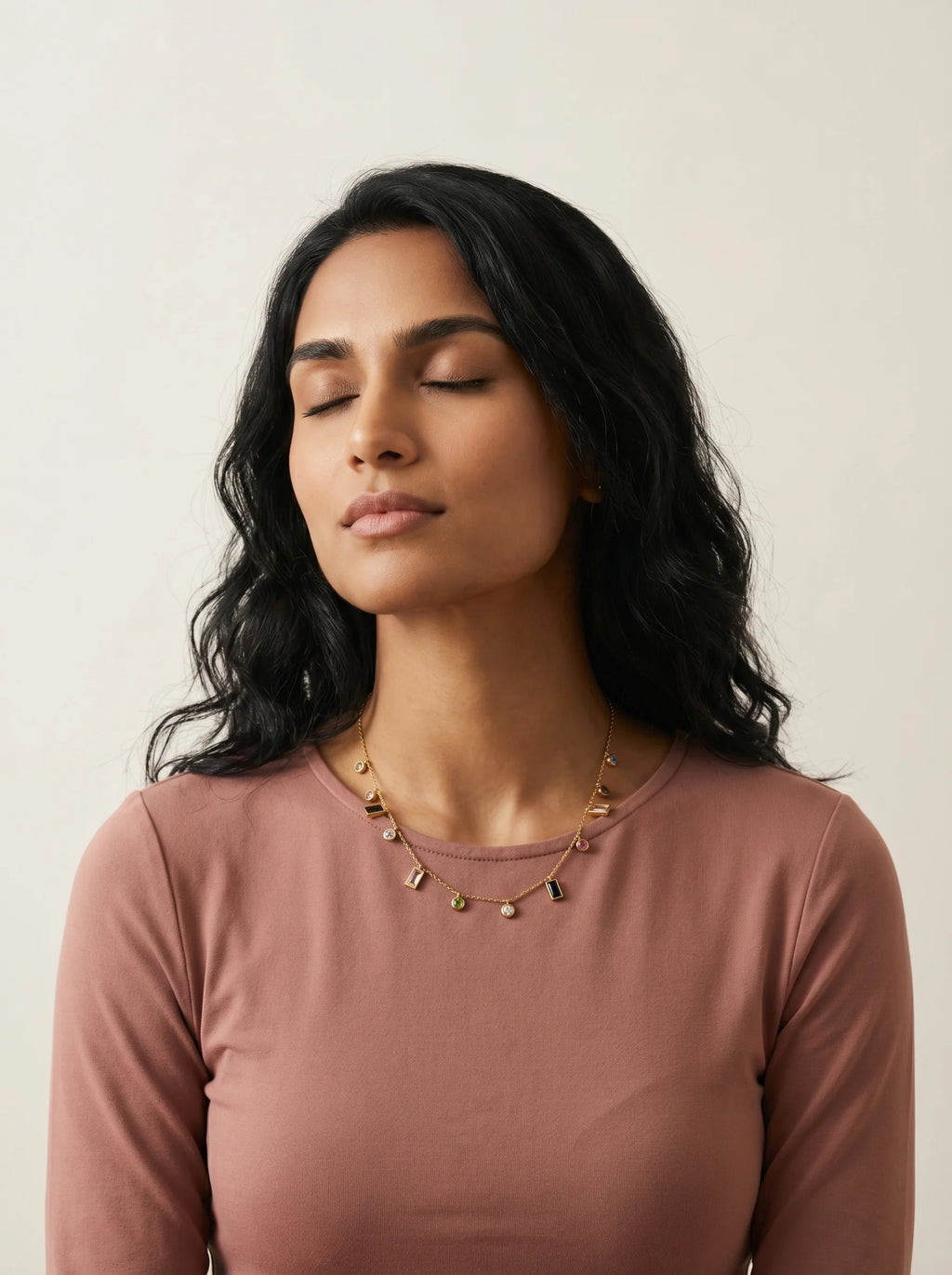 Aurum & Isle Mosaic gold charm necklace on model in dusty rose top, eyes closed — soft editorial moment.