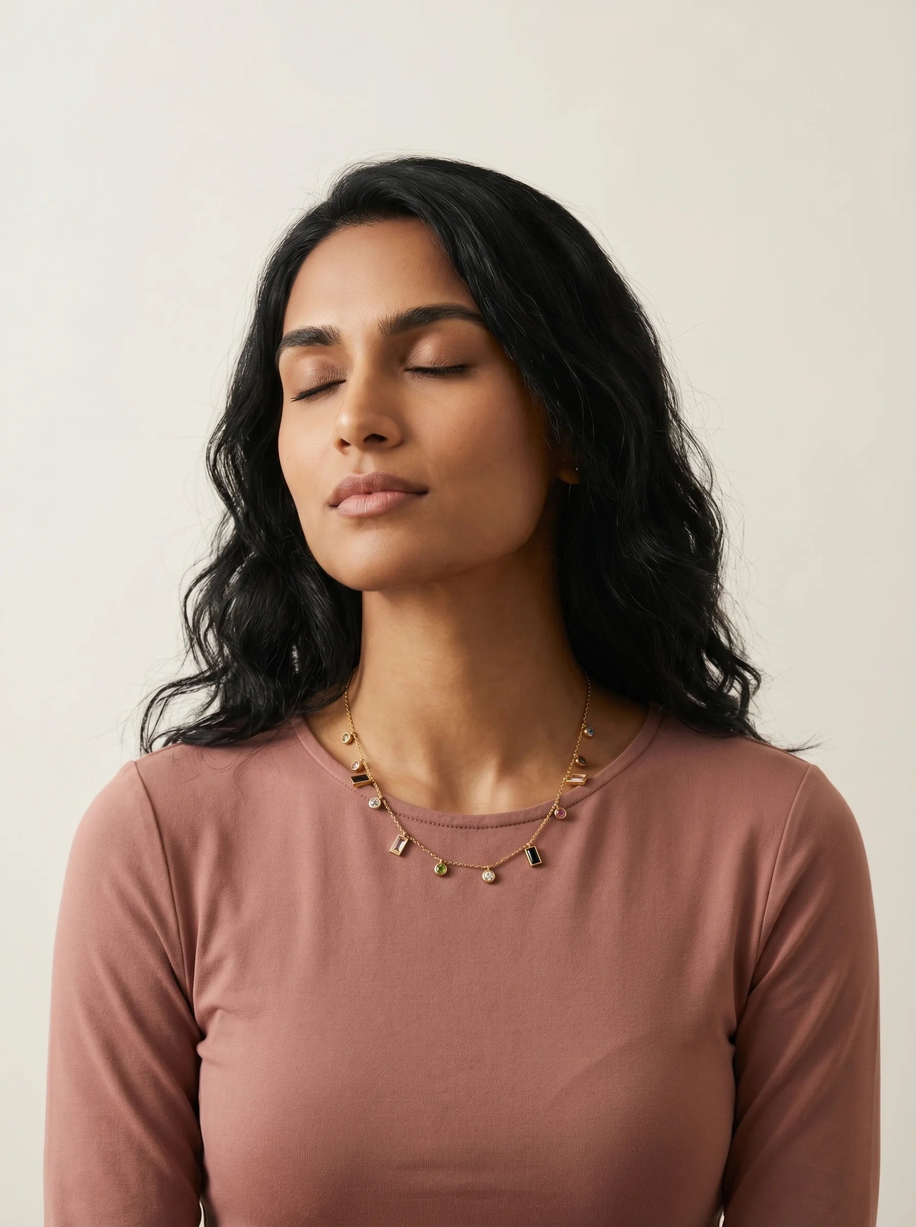 Aurum & Isle Mosaic gold charm necklace on model in dusty rose top, eyes closed — soft editorial moment.