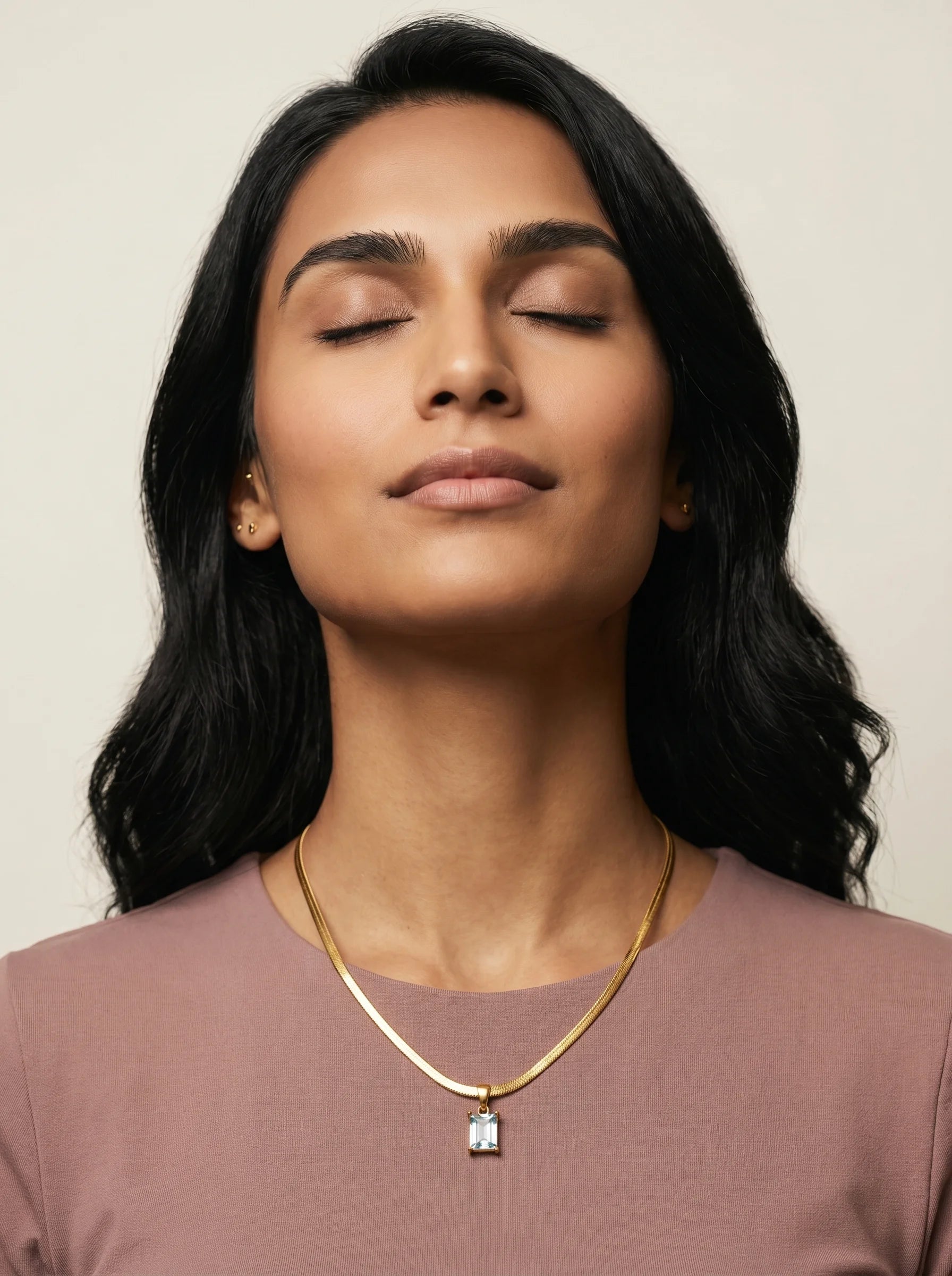 Aurum & Isle Soleil snake chain pendant on model in dusty rose top, eyes closed — soft editorial moment.