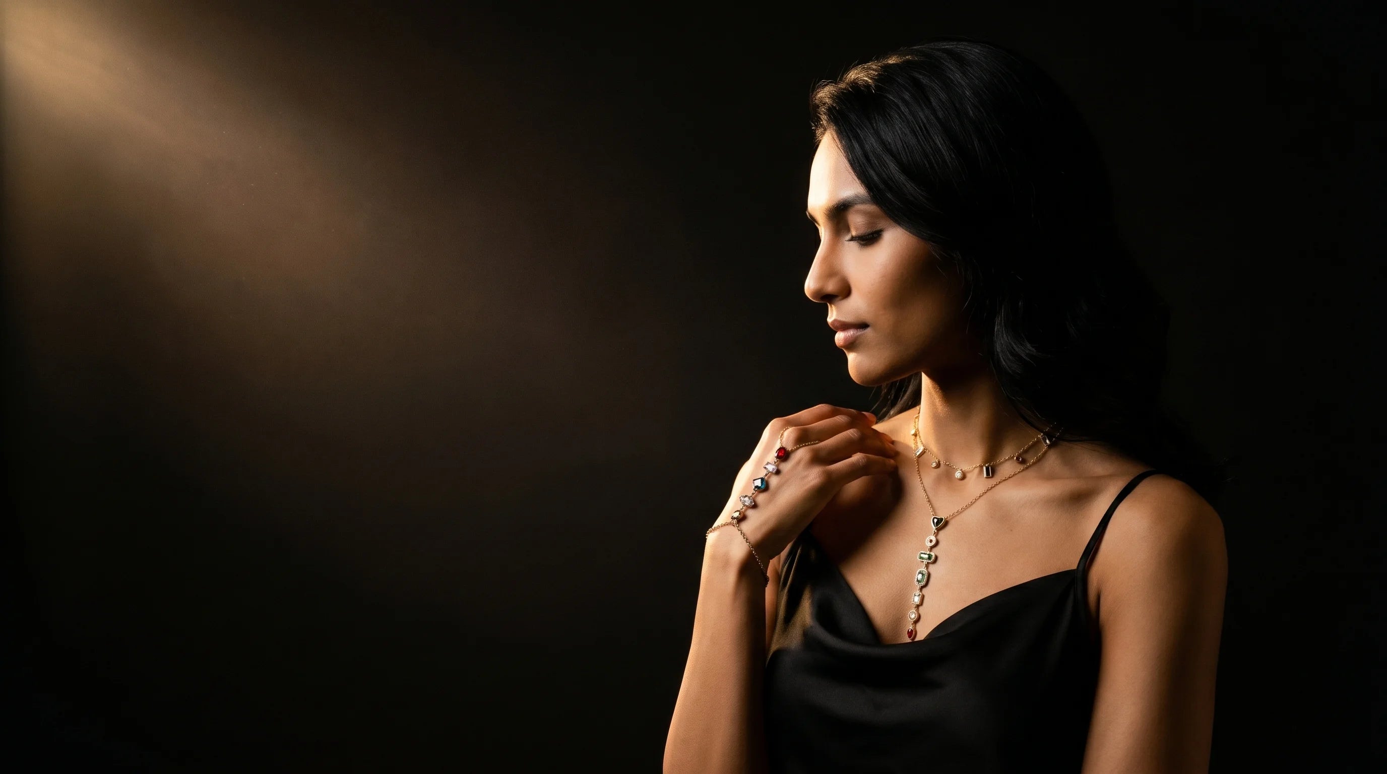 Cinematic moody editorial — woman in black slip wearing layered Aurum & Isle gold jewellery, lit by warm tungsten beam against deep black backdrop.